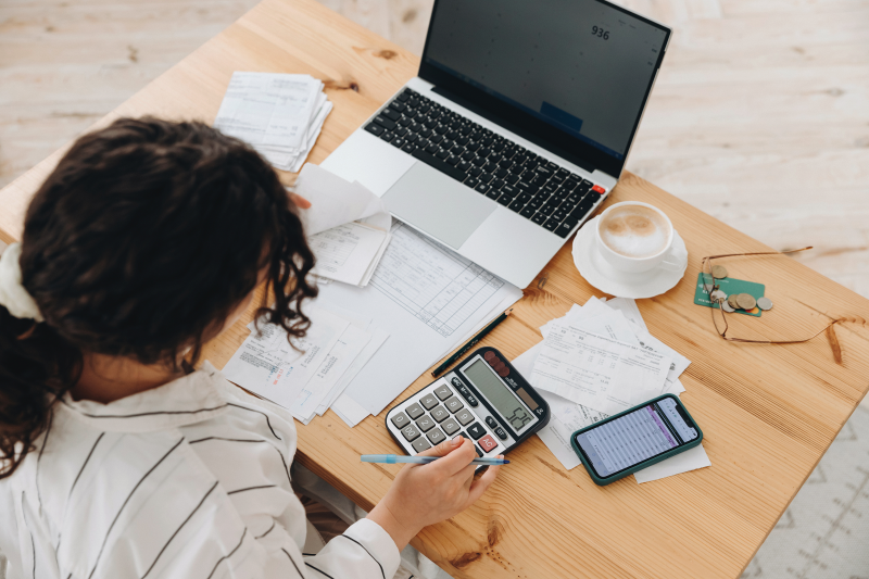 Student using a calculator and laptop with documents and receipts spread on the table