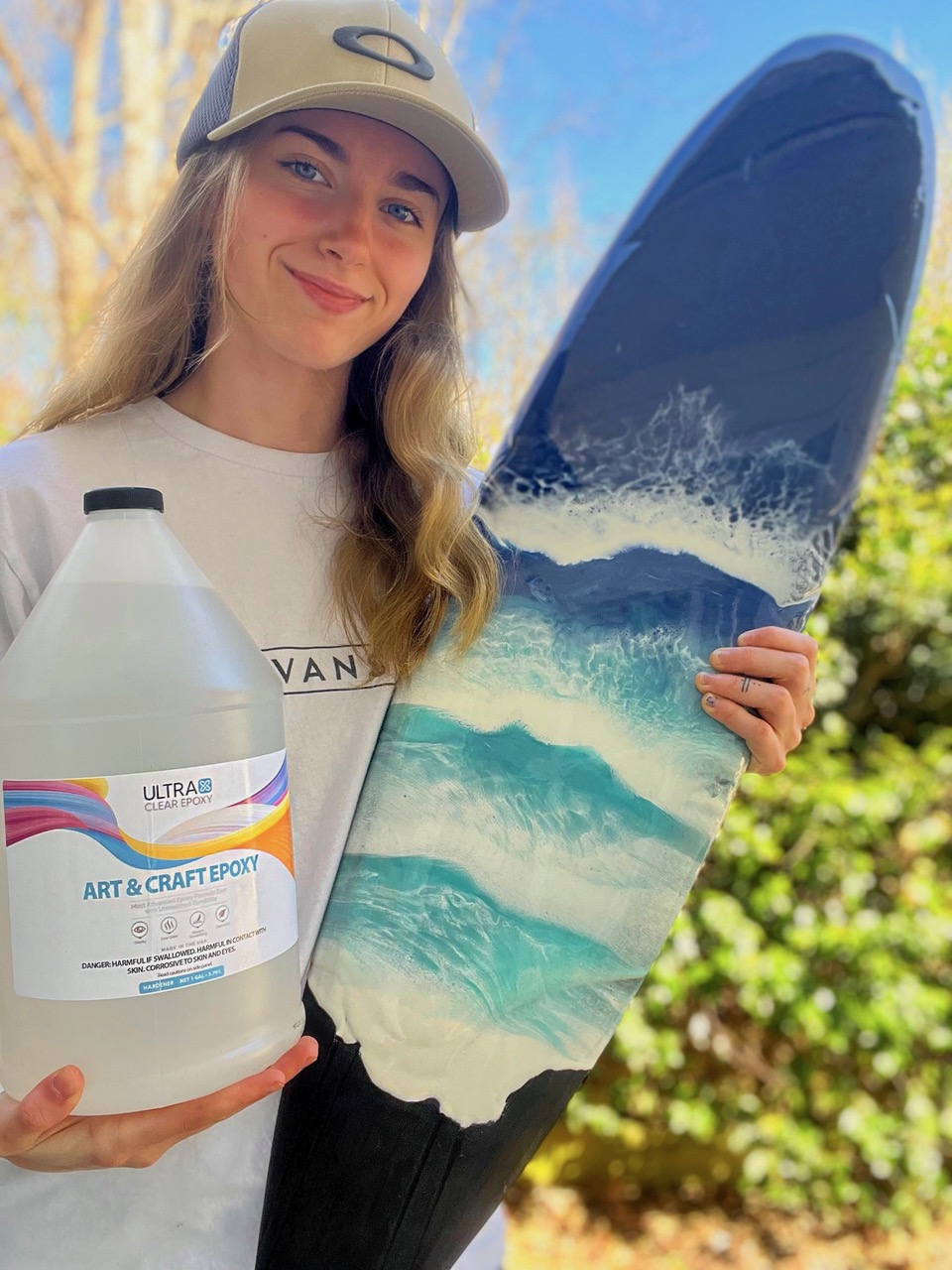 A woman holding a bottle of UltraClear Art & Craft Epoxy in her right hand and an epoxy art project made using it in her left hand. The art project consists of a surfboard-shaped piece of painted wood with several layers of epoxy that have each been pigmented a different blue or white tone using Pigmently powder pigments. The epoxy has been layered and manipulated to depict the motion of foamy ocean waves shoring up on a beach.