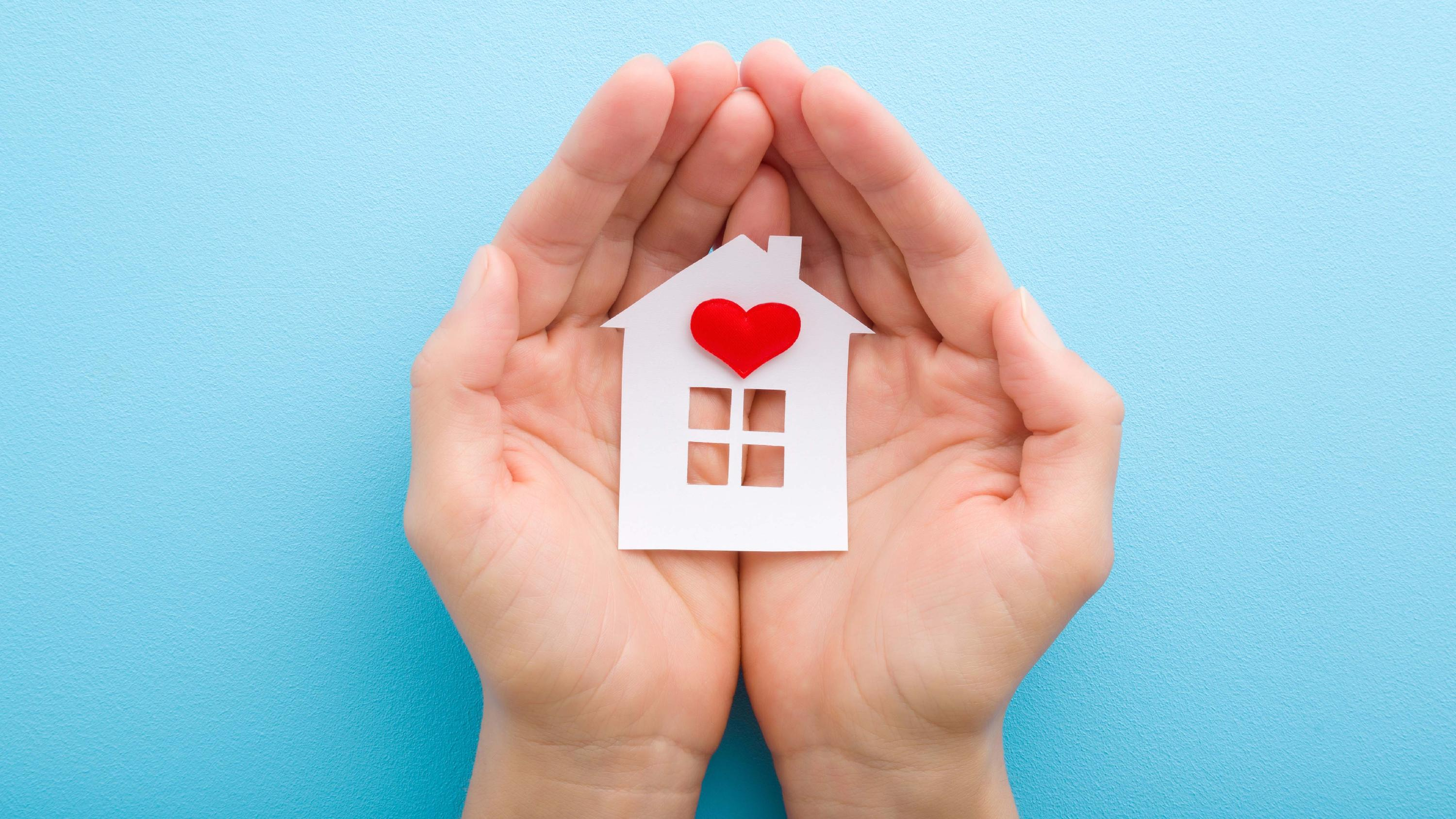 Two hands gently holding a paper house with a red heart symbol on its front, representing love and care for a home.