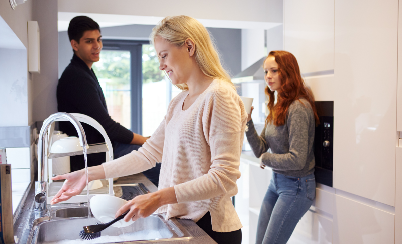 Three students hanging out in a kitchen while one washes dishes.