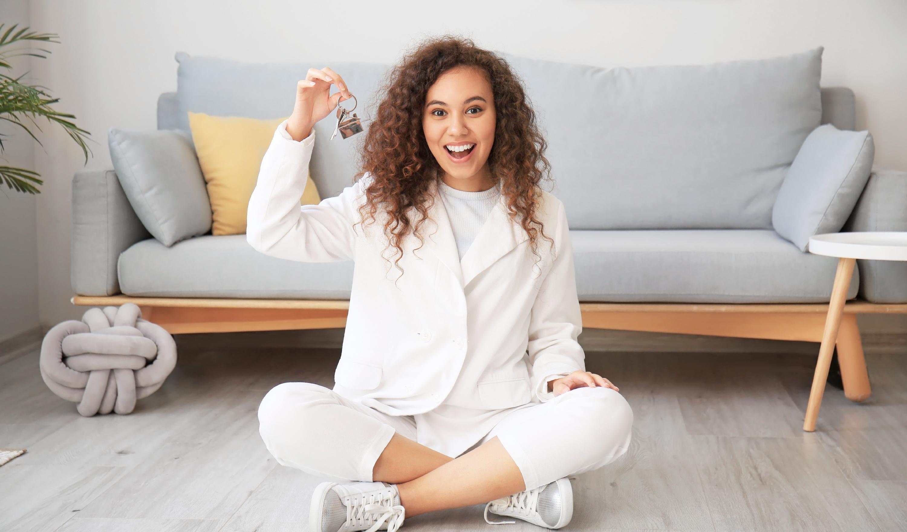 Student smiling and holding up keys while sitting on an apartment floor.