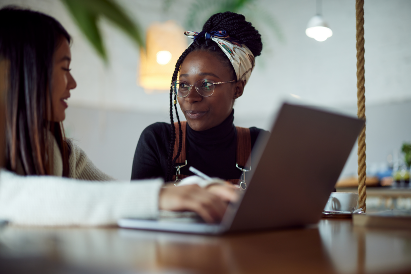 Two students seated at a table, using a laptop and talking together