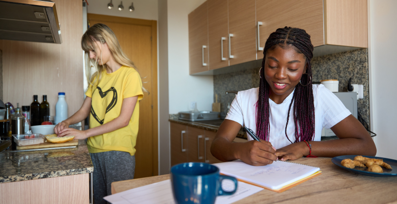 One roommate studying at the kitchen table while the other prepares food.